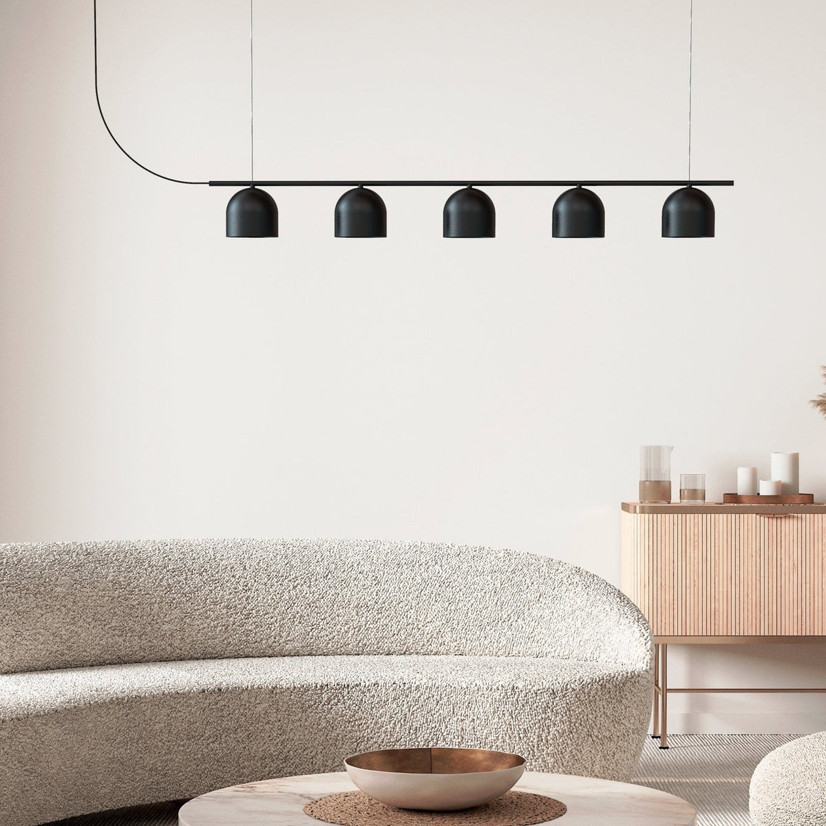 Rio Linear 5 Light Dome Chandelier in matt black iron, five small dome shades along a slim horizontal bar, shown above a boucle sofa in a living room setting.