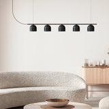 Rio Linear 5 Light Dome Chandelier in matt black iron, five small dome shades along a slim horizontal bar, shown above a boucle sofa in a living room setting.