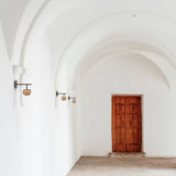 Roi single arm wall light with black backplate and arm, ribbed fumé glass dome shade, mounted in a white vaulted corridor near a wooden door.