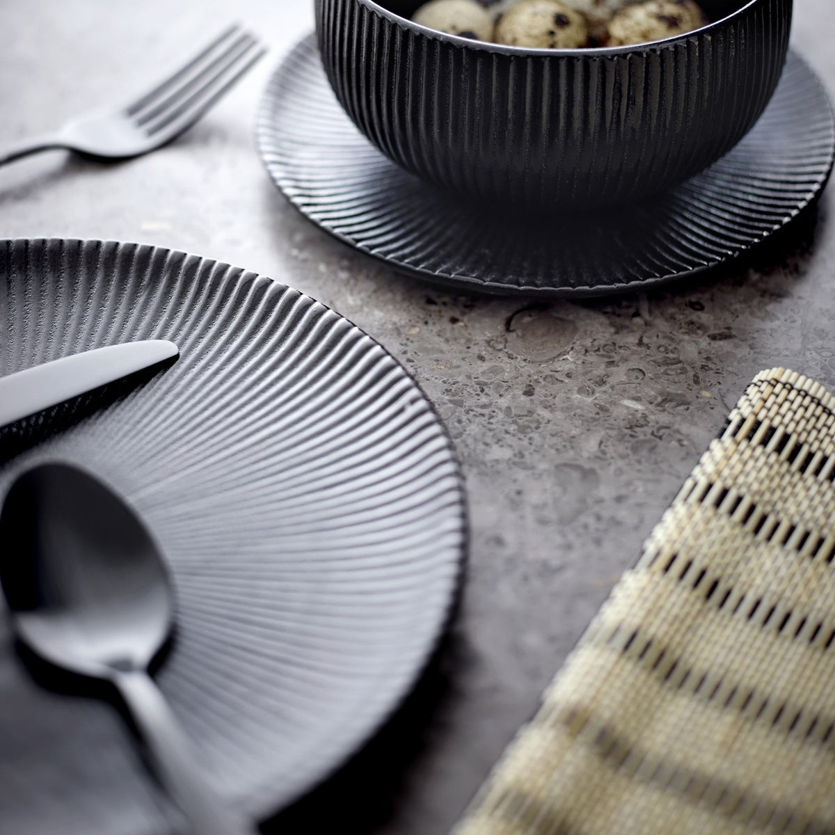 Neri Stoneware Side Plate in black with ribbed sunburst texture, set on a grey surface alongside cutlery and a matching ribbed bowl.
