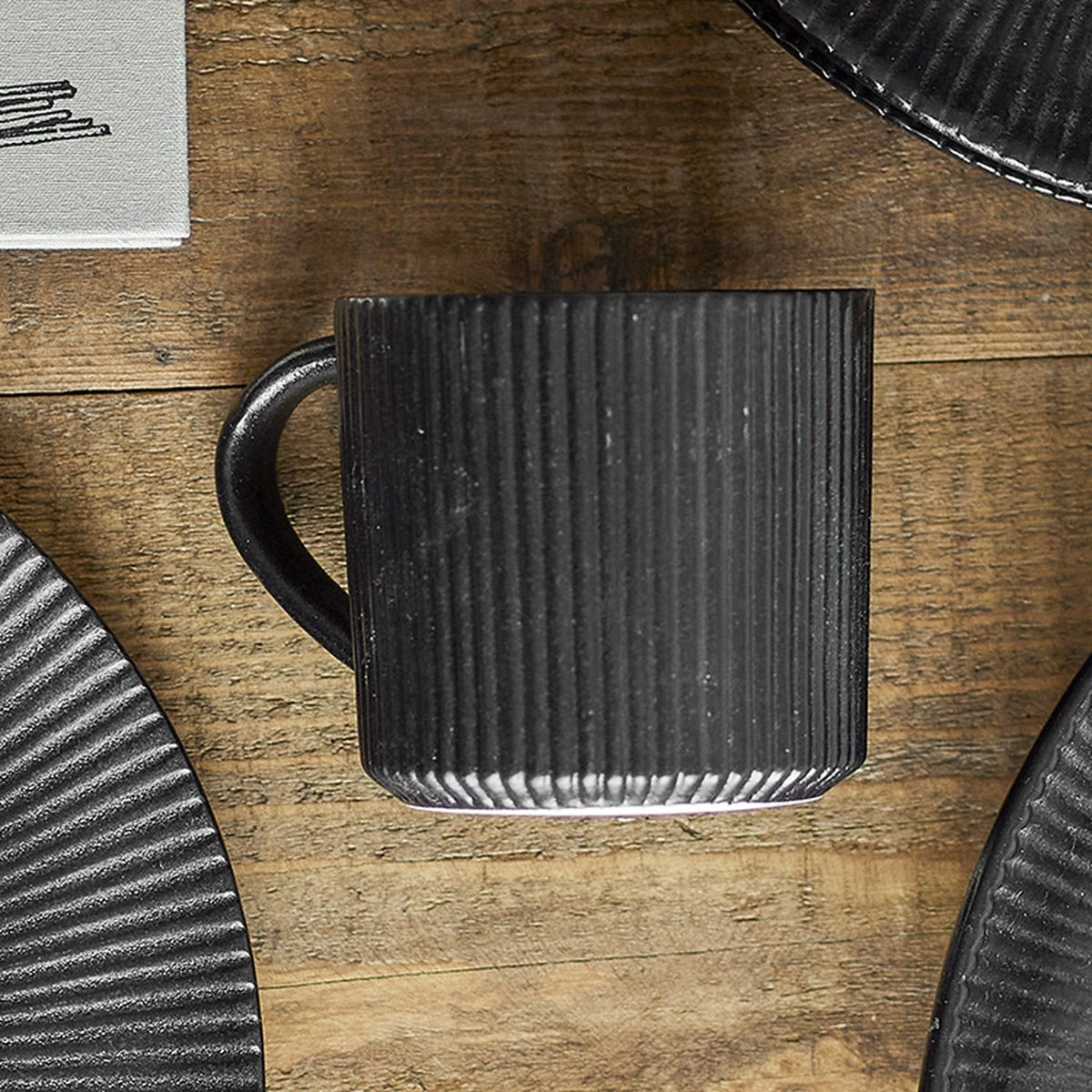 Neri Stoneware Cup 350ml in black, a ribbed ceramic mug with loop handle and smooth base band, shown on a wood surface beside matching dark plates.