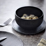 Neri Stoneware Bowl 14.5cm in black with vertical ribbed exterior, shown on a matching ribbed black plate alongside quail eggs on a grey stone surface.