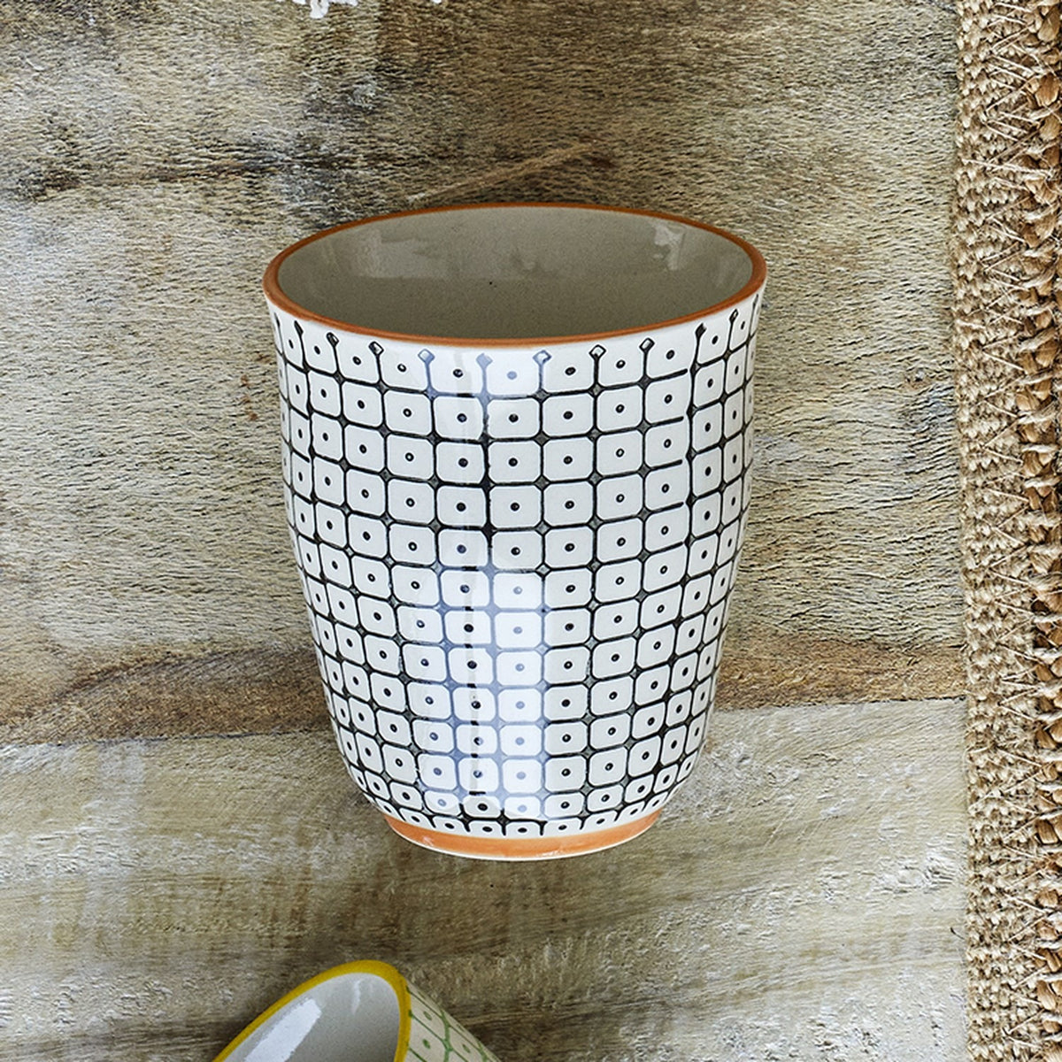 Carla Cup handleless stoneware tumbler in white with black grid-and-dot pattern and orange rim and base trim, shown on a wooden surface.