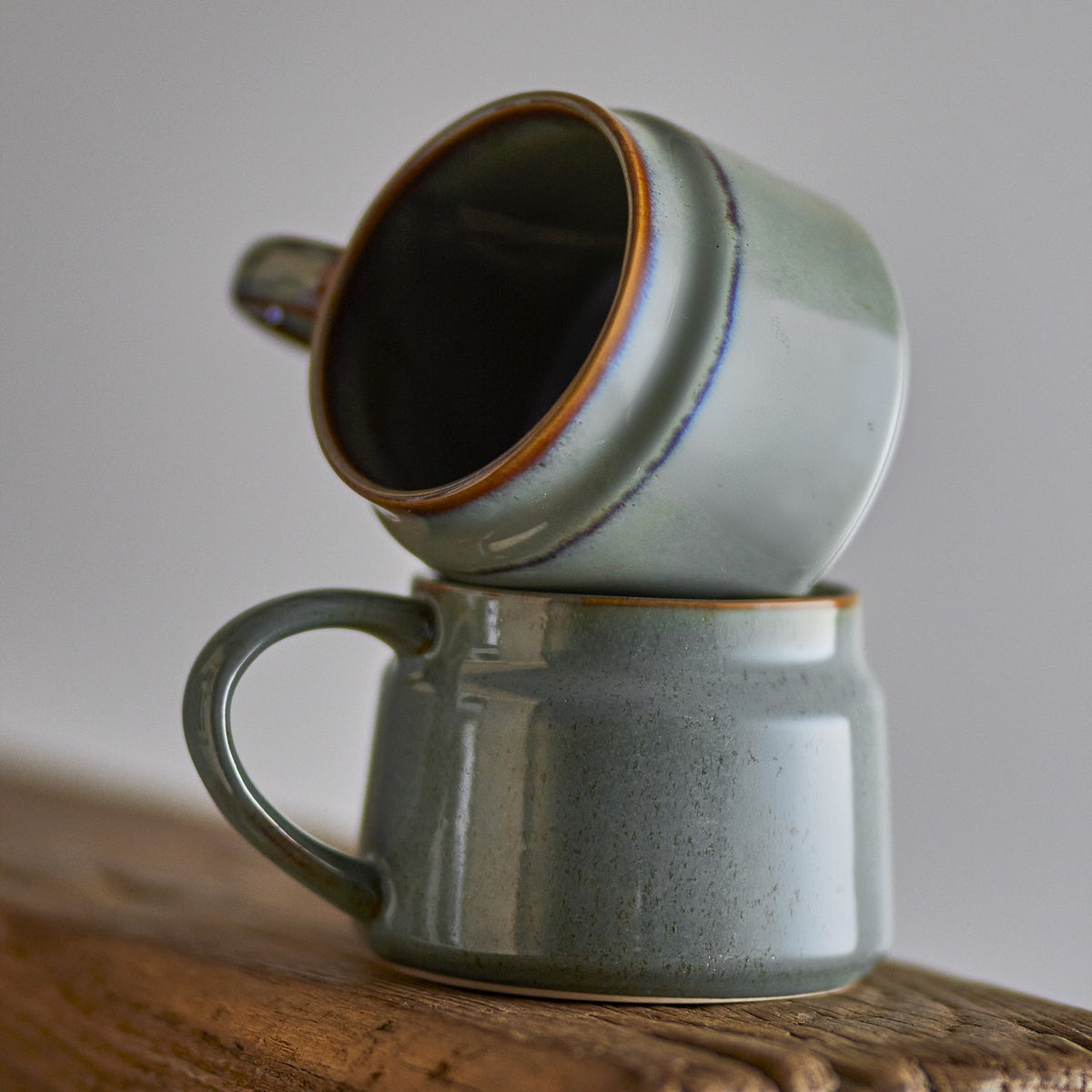 Two Pixie stoneware cups in green with open loop handles, one stacked tilted on top of the other, on a wooden surface against a plain grey background.