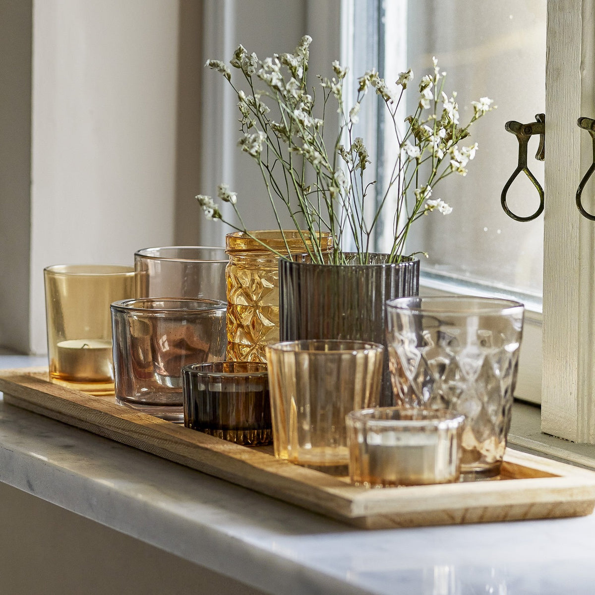 Sanga Glass Trays with Tealight Holders Set of 10 in brown, showing varied glass votives—plain, ribbed, faceted and diamond-patterned—on a wooden tray by a window with sprigs of gypsophila.