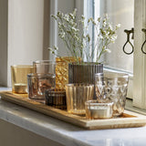 Sanga Glass Trays with Tealight Holders Set of 10 in brown, showing varied glass votives—plain, ribbed, faceted and diamond-patterned—on a wooden tray by a window with sprigs of gypsophila.