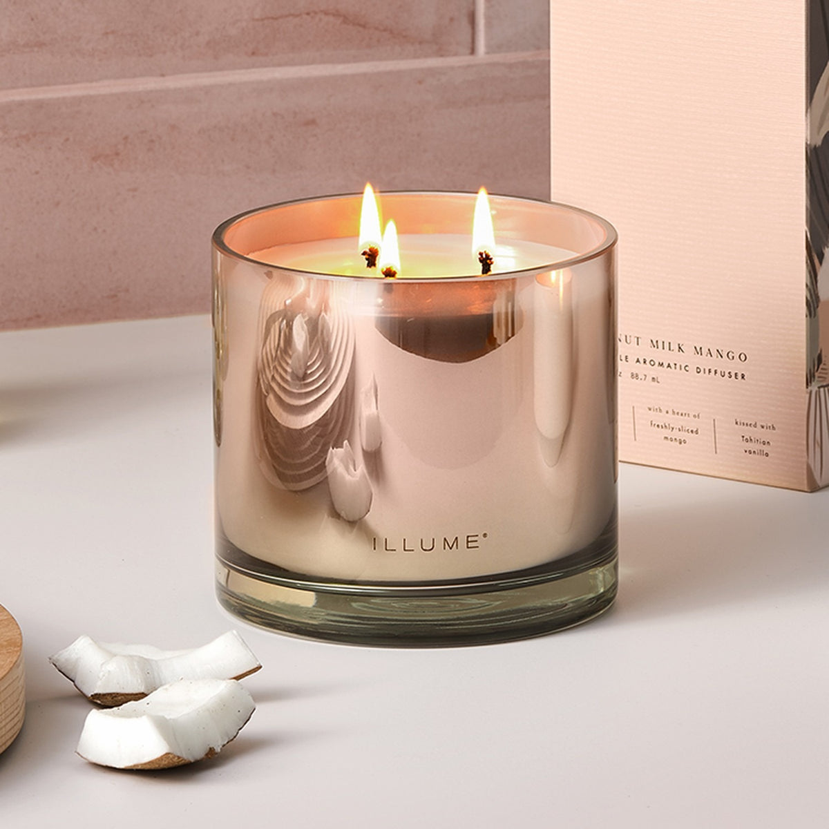 Coconut Milk Mango scented candle in a pink ombre glass vessel with three lit cotton wicks, shown beside coconut pieces and a gift box on a light surface.