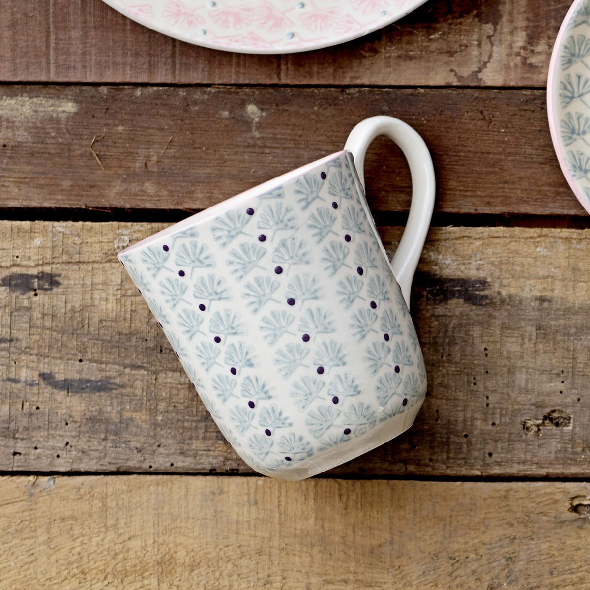 Maya Mug with white glaze, pale blue floral leaf pattern and small dark dots, curved white handle, shown on a wooden surface beside matching patterned plates.
