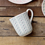Maya Mug with white glaze, pale blue floral leaf pattern and small dark dots, curved white handle, shown on a wooden surface beside matching patterned plates.