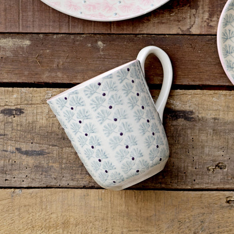 Maya Mug with white glaze, pale blue floral leaf pattern and small dark dots, curved white handle, shown on a wooden surface beside matching patterned plates.