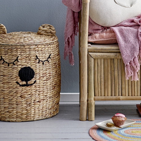 Dolma Bear Water Hyacinth Basket with Lid in natural woven finish, featuring stitched sleepy bear face and rounded ears, shown in a room beside a bamboo bench with pink throws.