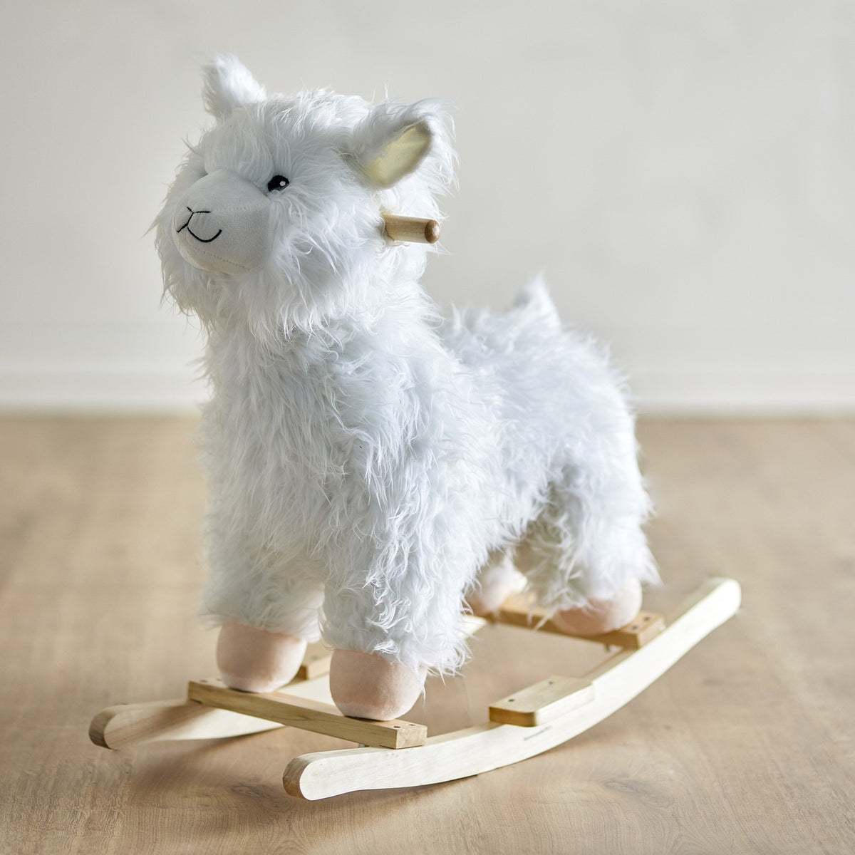 Kinto Rocking Llama in white, a shaggy fabric ride-on toy with embroidered face, wooden handle grip, and curved wooden rockers, shown on a light wood floor.