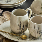 Bea Crackle Stoneware Mug in cream with botanical stem print, rounded body and curved handle, shown on a matching patterned plate with a gold spoon and a second mug.