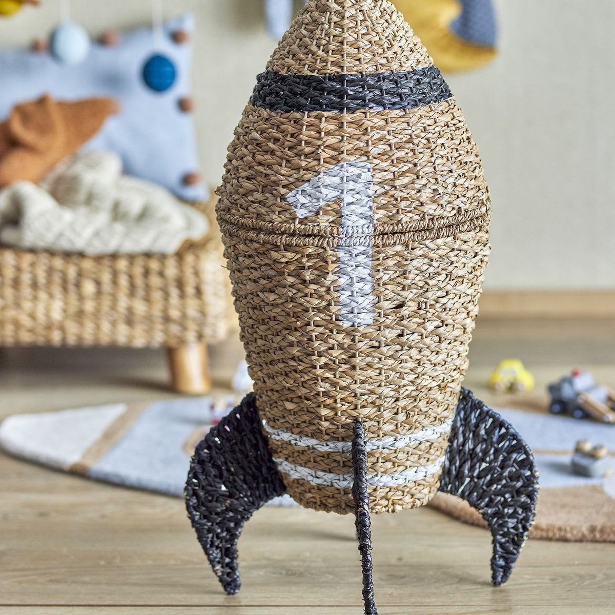 Cosmina Rocket Seagrass Basket with Lid in natural weave with dark band and "1" motif, fins, and lid, shown in a nursery setting.