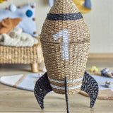 Cosmina Rocket Seagrass Basket with Lid in natural weave with dark band and "1" motif, fins, and lid, shown in a nursery setting.