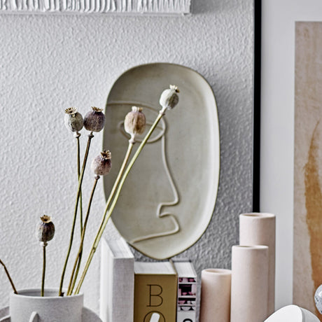 Ngan Stoneware Tray in muted green, an oval ceramic tray with a raised line-drawn face design, displayed on a shelf with dried poppy seed heads and books.
