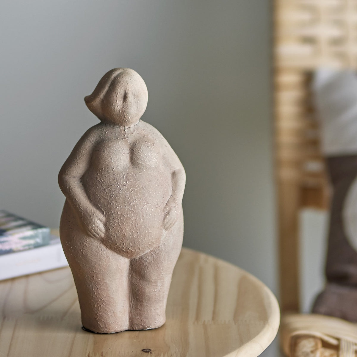 Sidsel Terracotta Ornament in brown, a standing female figurine with rounded curves, hands resting on her belly and swept side hair, shown on a wooden table.