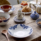 Camellia Porcelain Plate in cream with hand-painted blue leaf border, shown as a side plate on a rustic wooden table styled with bowls, a jug, teapot and bread for breakfast.