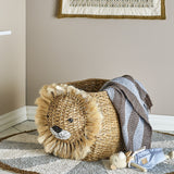 Harrison Basket in beige woven bankuan grass with a fringed lion head design, shown in a nursery holding a blanket and soft toy on a rug.