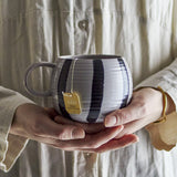 Serina Striped Stoneware Mug in grey with broad vertical stripes and ridged texture, held in both hands against a neutral beige background.