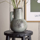 Tias Ceramic Vase 23cm in Grey with a round body, tall straight neck and flat handle, mottled finish, holding dried grasses on a black wooden stool indoors.