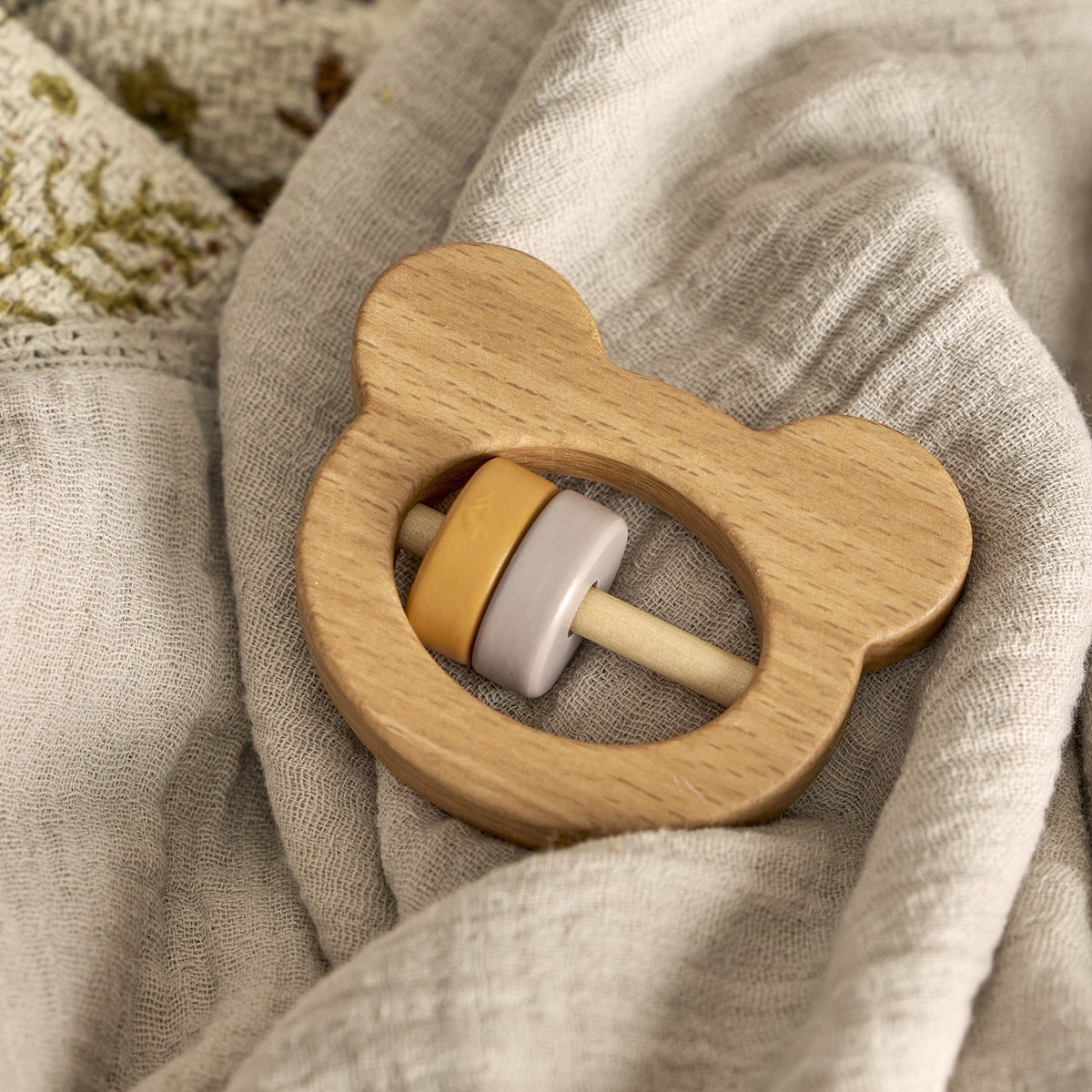 Wooden bear-shaped rattle from the Riina Baby Play Set, with mustard and grey ring beads, resting on a soft neutral fabric background.