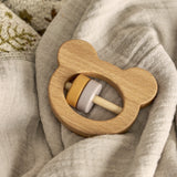 Wooden bear-shaped rattle from the Riina Baby Play Set, with mustard and grey ring beads, resting on a soft neutral fabric background.