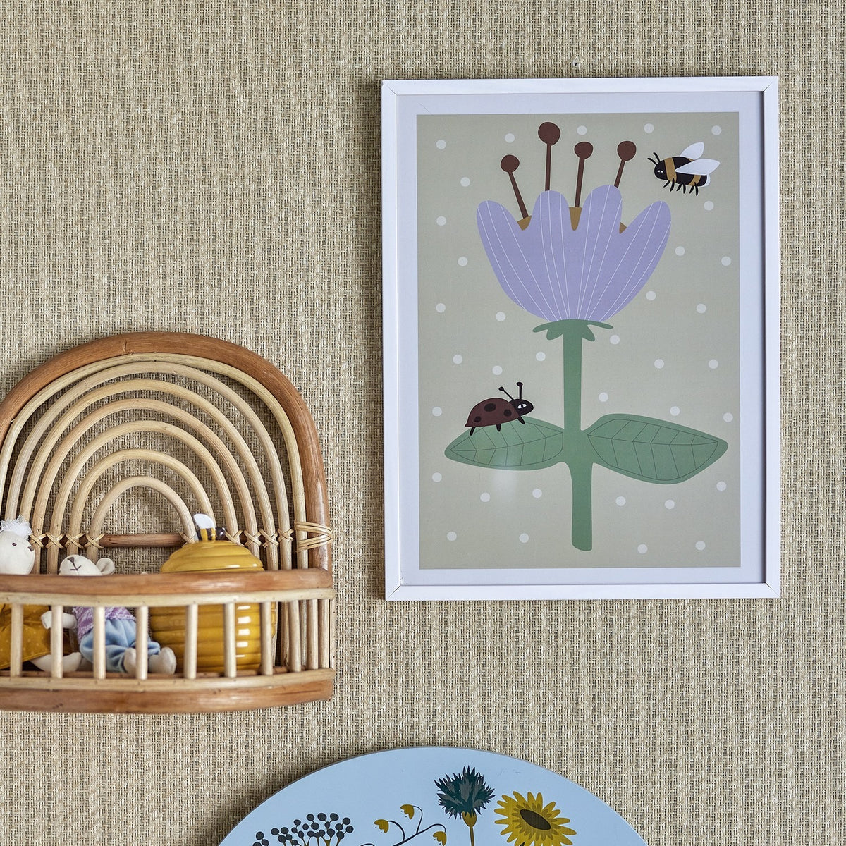 Numa Wooden Framed Print in white frame, showing a purple flower with stamens, bee, and ladybird on green leaves against a spotted sage background, hung on a woven wall near a rattan shelf.
