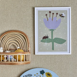 Numa Wooden Framed Print in white frame, showing a purple flower with stamens, bee, and ladybird on green leaves against a spotted sage background, hung on a woven wall near a rattan shelf.