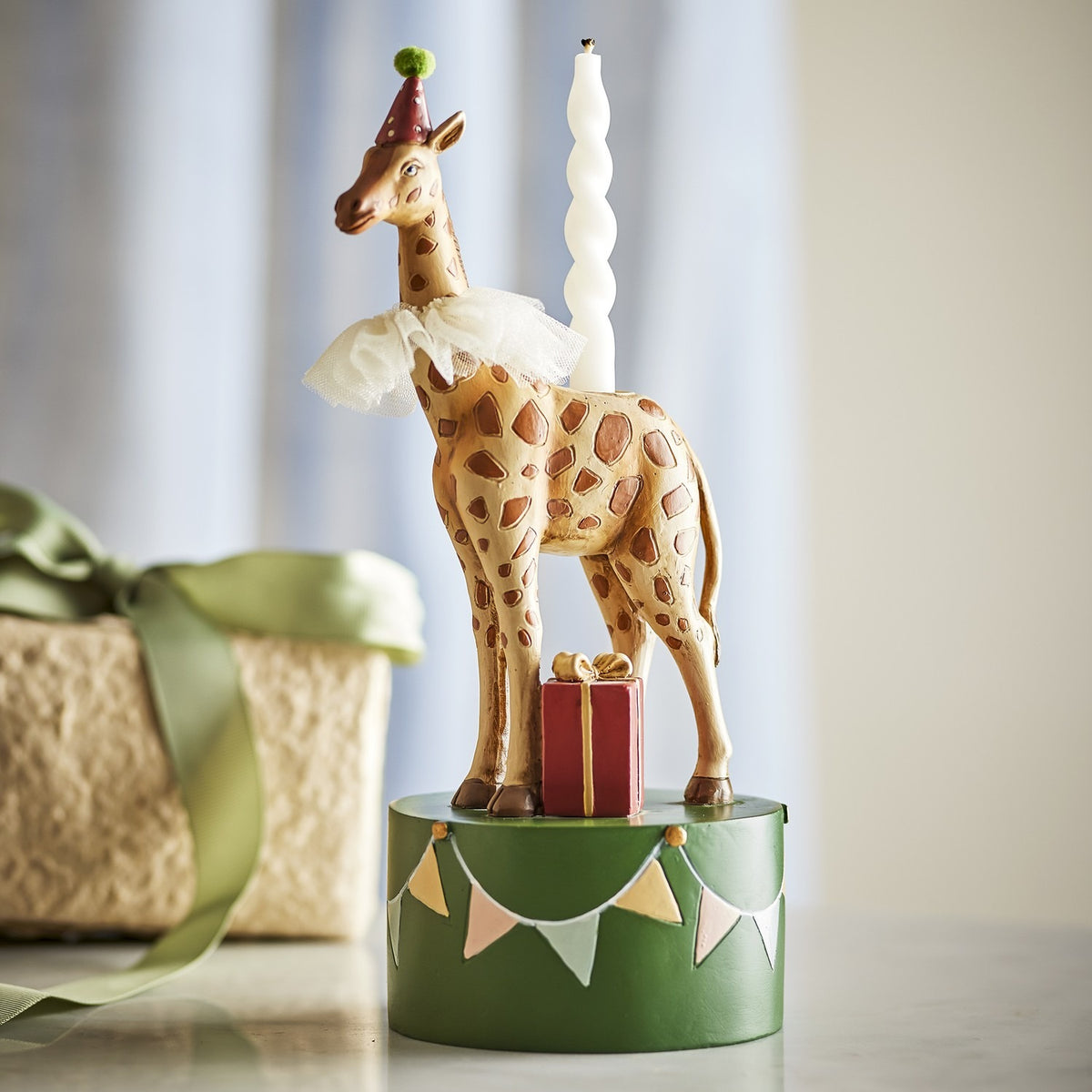Flor resin candle holder, 25cm, shaped as a giraffe in a party hat and frilled ruff on a green bunting-painted base, with a spiral candle and small wrapped gift, set against a soft neutral backdrop.
