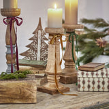 Noore Wooden Candle Holder 31cm in natural mango wood with a turned column and ridged collars, shown holding a lit pillar candle tied with a gold ribbon amid festive décor.