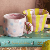 Nini Dotted Stoneware Cup in pink with blue dot pattern and red striped ring handle, shown beside a yellow striped mug on a book.
