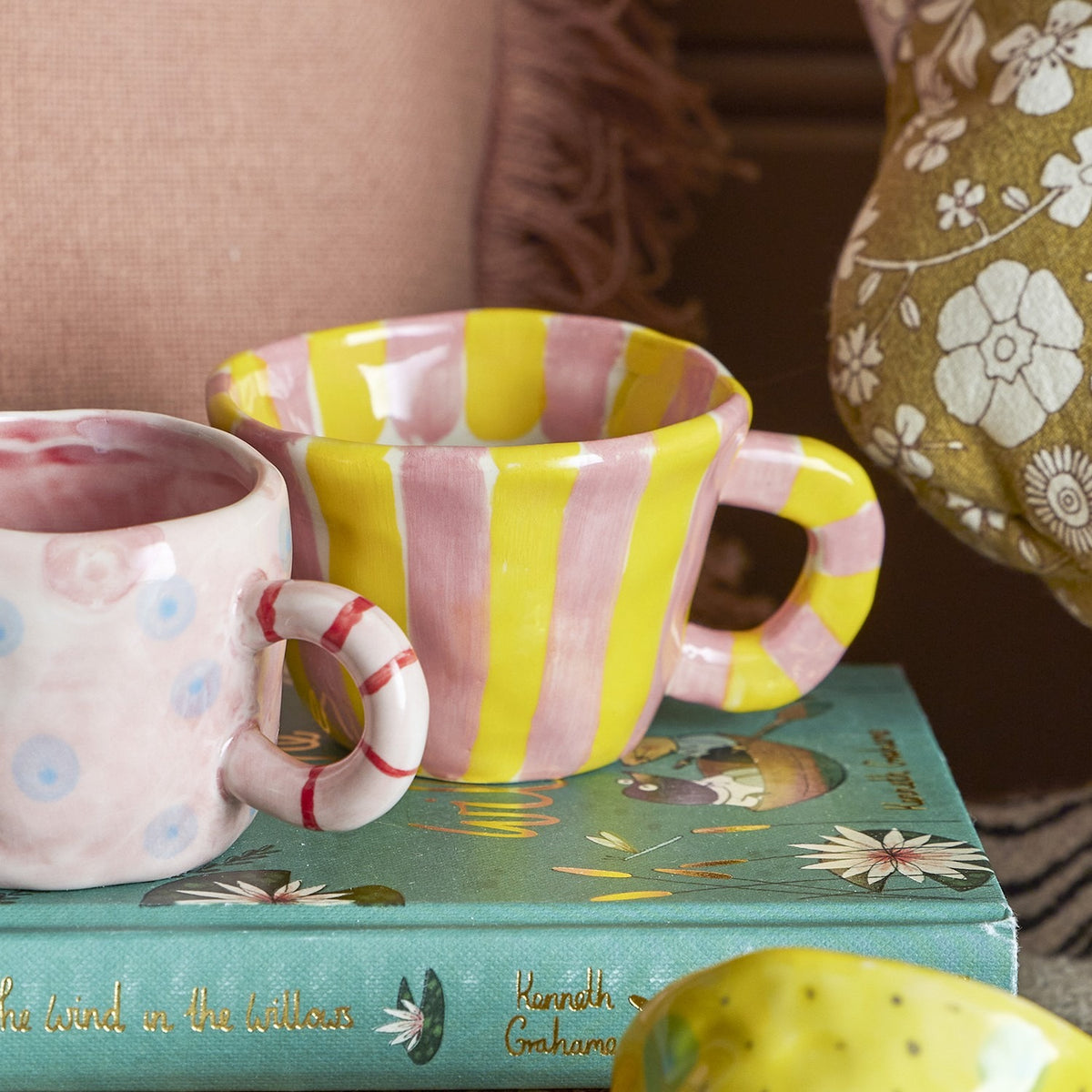 Nini Yellow Striped Stoneware Cup 110ml in pink and yellow with a loop handle, shown beside a polka-dot cup atop a book in a living room setting.