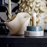 Brent Stoneware Candle Holder in beige, a sculptural curved piece with speckled sandy finish, shown holding a twisted taper candle beside a marble pillar candle stand.