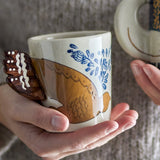 Hezha Stoneware Cup in cream, hand-painted with a bird in flight and blue floral sprigs, its fanned tail forming the handle, held in two hands.