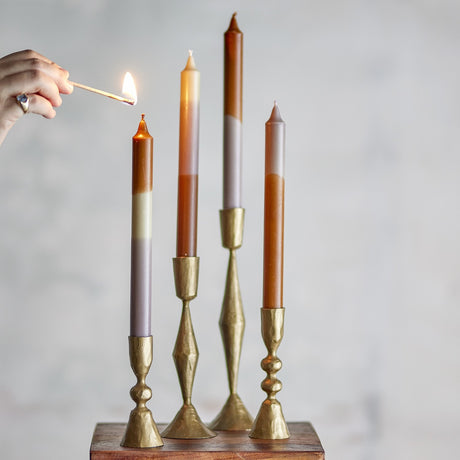 Dip Dye Fluted Candles Pack of 4 in brass candlesticks against a light grey background, each slim taper showing multicoloured dip-dye bands as a hand lights one with a match.