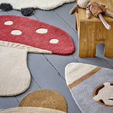 Mushroom Wool Rug 90x85cm in red, shaped like a toadstool with white spots and cream stalk, shown on grey floorboards beside other kids' rugs and toys.