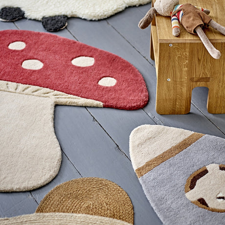 Mushroom Wool Rug 90x85cm in red, shaped like a toadstool with white spots and cream stalk, shown on grey floorboards beside other kids' rugs and toys.