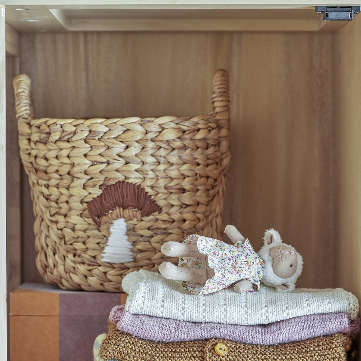 Agnes Water Hyacinth Basket with handles and stitched mushroom motif in beige, shown on a shelf with folded knits and a toy lamb.