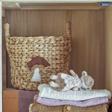 Agnes Water Hyacinth Basket with handles and stitched mushroom motif in beige, shown on a shelf with folded knits and a toy lamb.