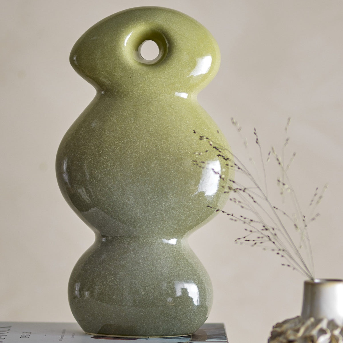 Kaui Stoneware Ornament in green, a sculptural stack of rounded ceramic forms with a pierced oval head, glossy speckled glaze shading from light to deep green, set against a neutral background.