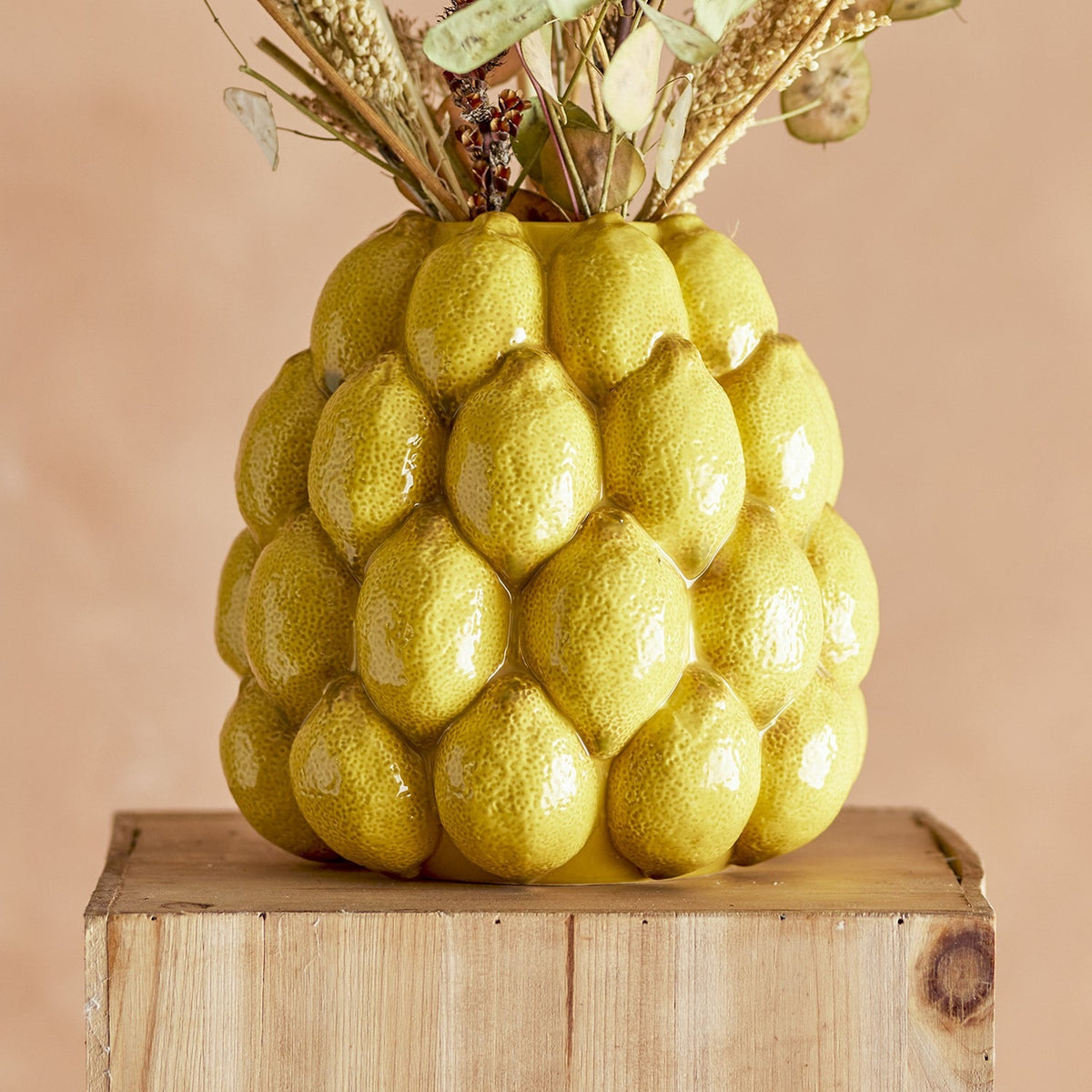 Limone Stoneware Vase 22cm in yellow, moulded as clustered glossy lemons with a narrow round opening, holding dried flower stems on a wooden block against a peach background.