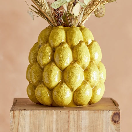 Limone Stoneware Vase 22cm in yellow, moulded as clustered glossy lemons with a narrow round opening, holding dried flower stems on a wooden block against a peach background.