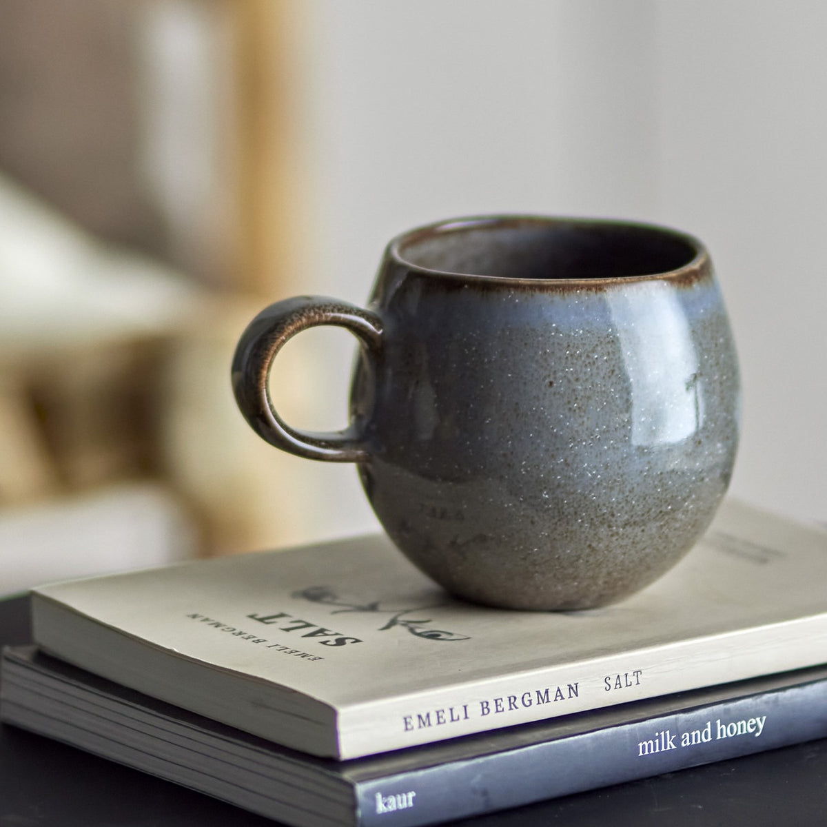 Sandrine Stoneware Cup 275ml in grey with a round, ball-like body and loop handle, speckled reactive glaze darkening towards the rim, resting on stacked books.
