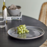 Sandrine Stoneware Dinner Plate in grey with speckled reactive glaze and raised rim, shown with green grapes on a dark table in a dining setting.
