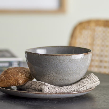 Sandrine Stoneware Bowl 15cm in grey with speckled reactive glaze and rounded sides, shown on a matching plate beside a bread roll and knitted napkin.