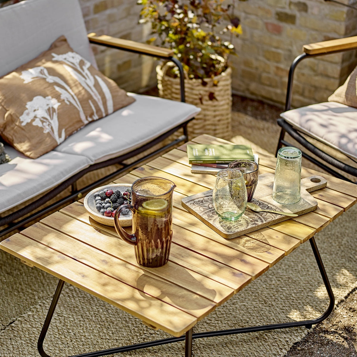 Hampton Wooden Garden Table in brown with slatted acacia top and black metal legs, styled on a patio with drinks, a tray and cushioned chairs.