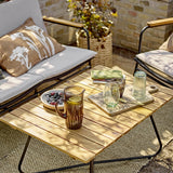 Hampton Wooden Garden Table in brown with slatted acacia top and black metal legs, styled on a patio with drinks, a tray and cushioned chairs.