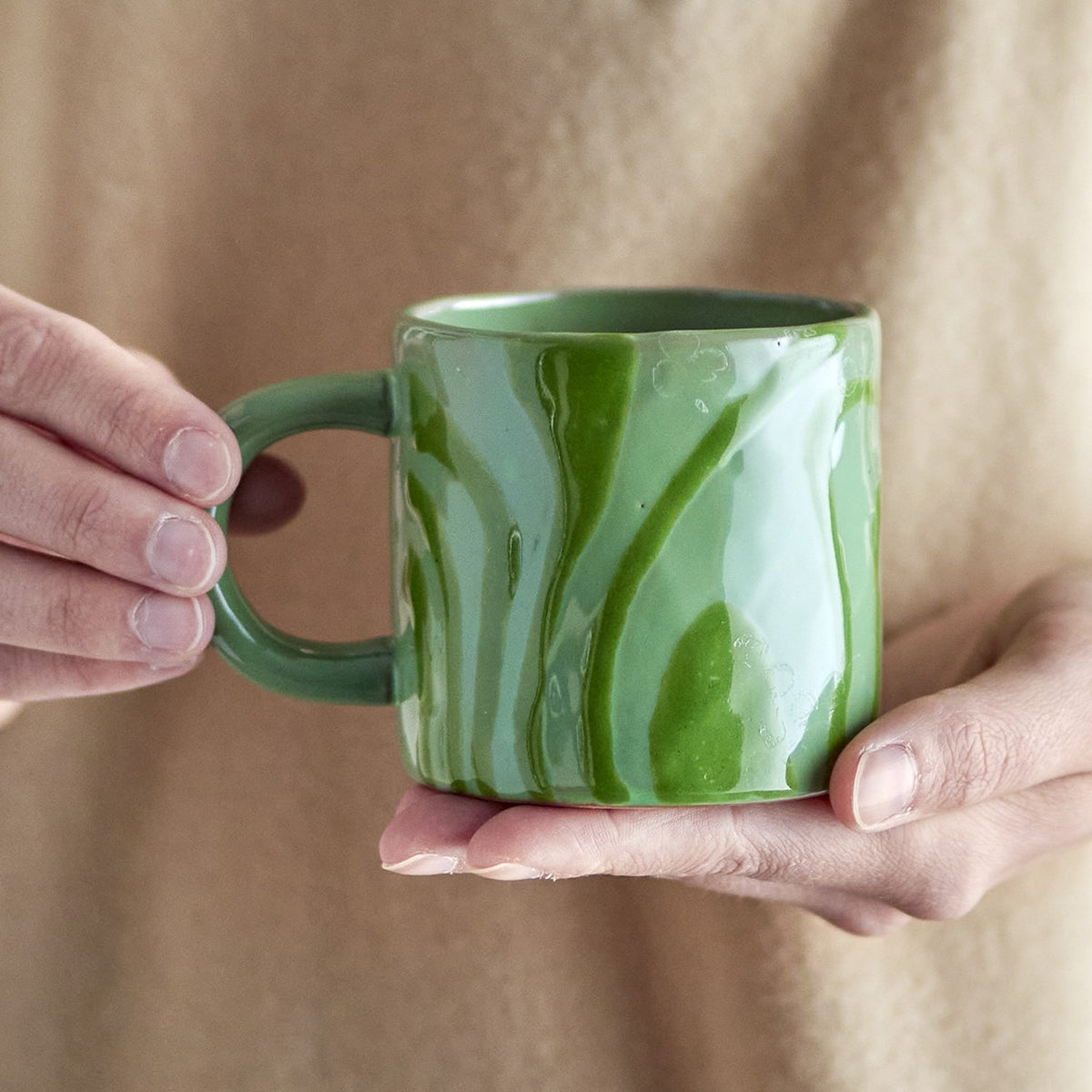 Ninka Stoneware Cup 200ml in Green, held in hand, showing the rounded loop handle and glossy glaze with bold flowing stripes from rim to base.
