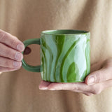Ninka Stoneware Cup 200ml in Green, held in hand, showing the rounded loop handle and glossy glaze with bold flowing stripes from rim to base.